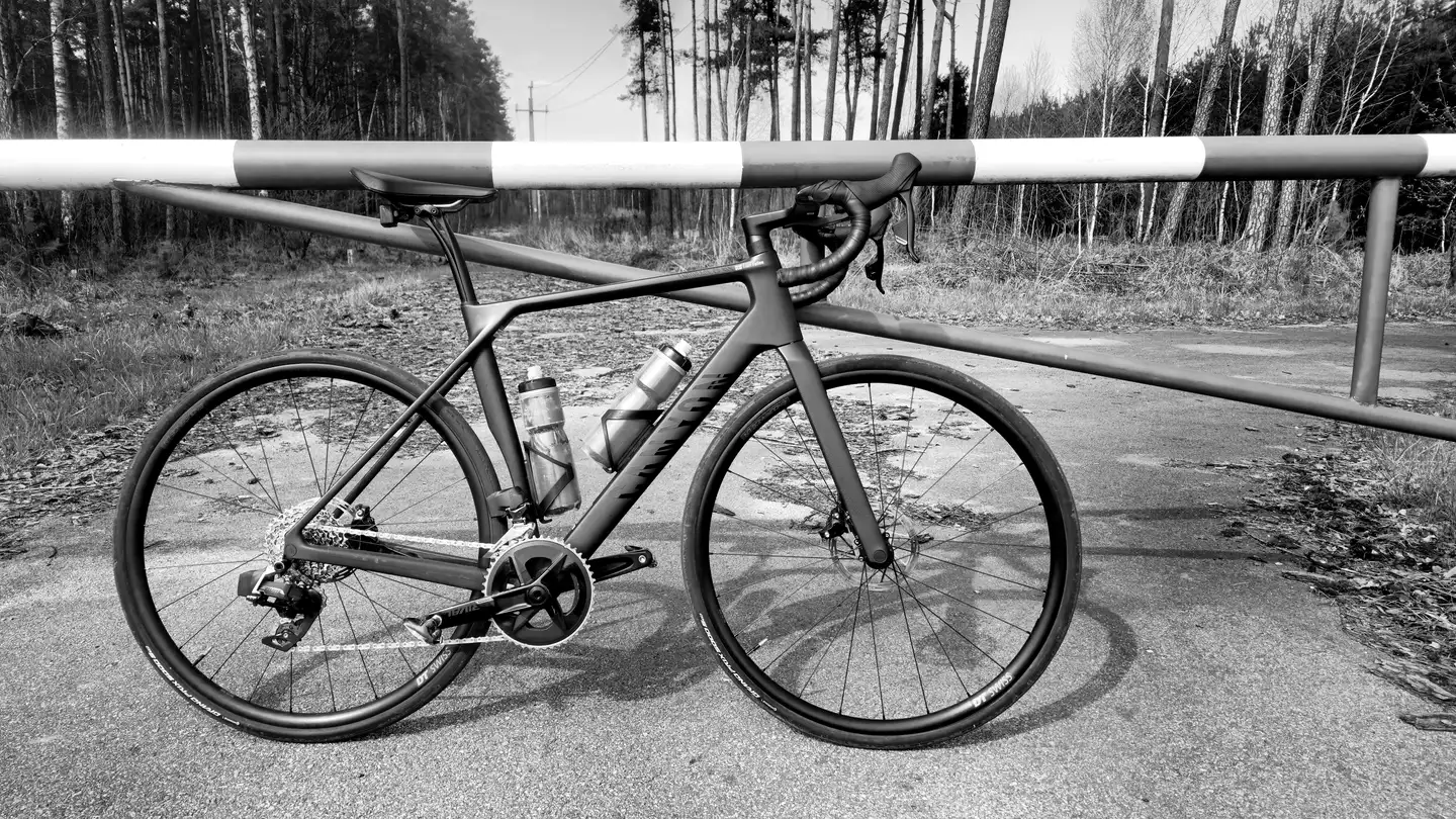 A black road bike propped against a barrier on an empty road lined with trees.