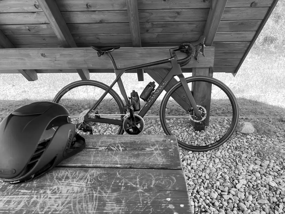 A road bike resting under a wooden shelter, with a helmet on the table in front.